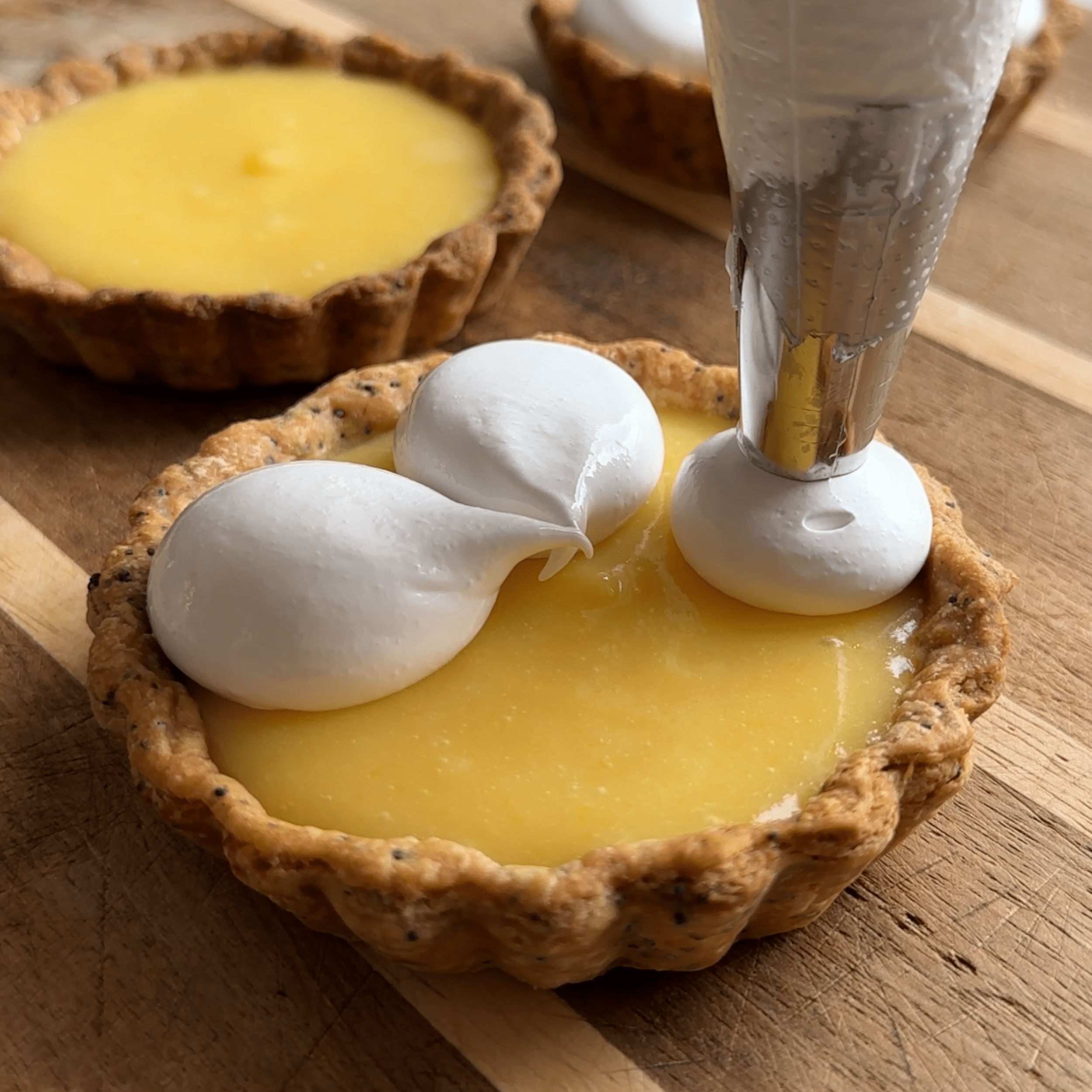 A wooden cutting board with 3 lemon tartlets. A circle piping tip is being used and midway of piping the 3rd petal of a 5 petal flower