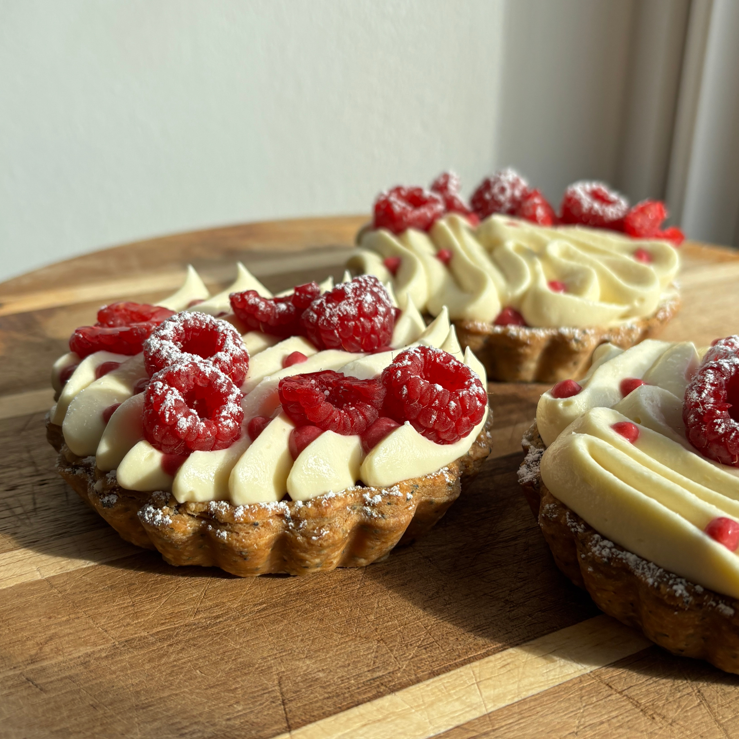 Three small raspberry tartlets on a wooden board in warm natural sunlight, topped with white chocolate whipped ganache, fresh raspberries dusted with powdered sugar. The foreground tart is in sharp focus, while the others are softly blurred in the background.